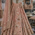 Blush pink embroidered lehenga with floral detailing, embellished borders, matching dupatta, and heavily decorated blouse displayed in a garment workshop
