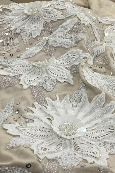 Close-up of white floral embroidery with sequins, beads, and intricate leaf detailing on sheer beige fabric